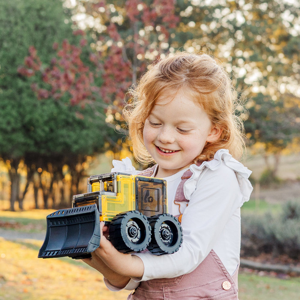 Young girl holding a toy bulldozer outdoors with trees in the background