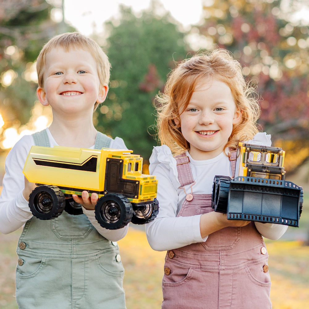 Two children holding magnetic tile toy construction vehicles outdoors.
