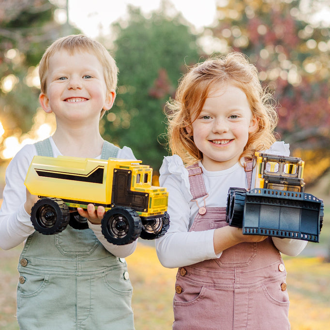 Two children holding magnetic tile toy construction vehicles outdoors.