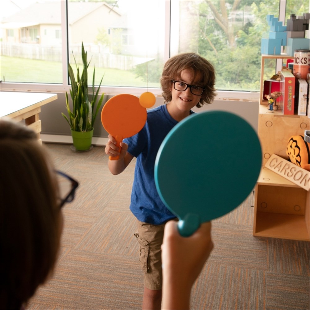 Two children playing with colourful paddles in a room with large windows and furniture.