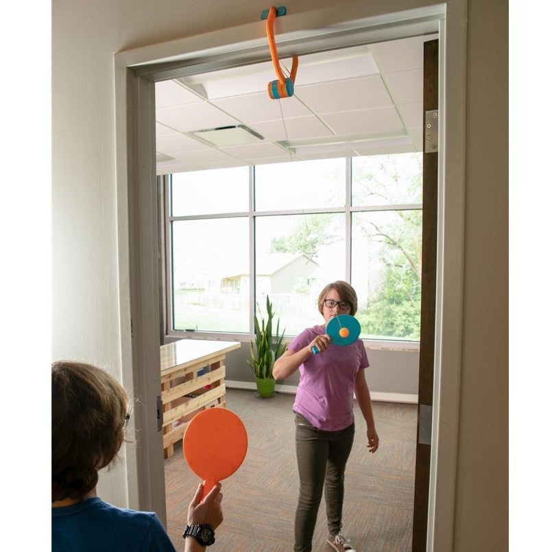 Two children playing with colorful paddles in a room with large windows.