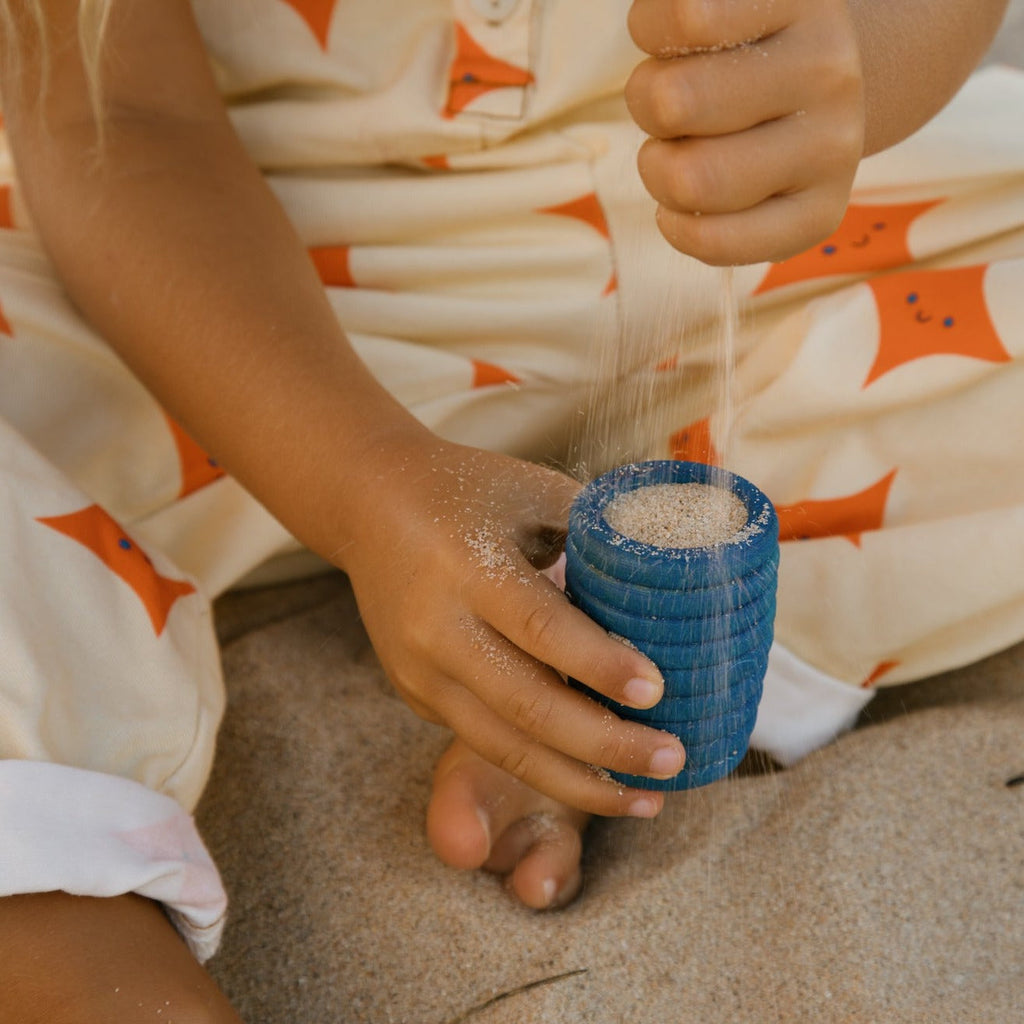 Child dropping sand into blue Grapat tall wooden pot