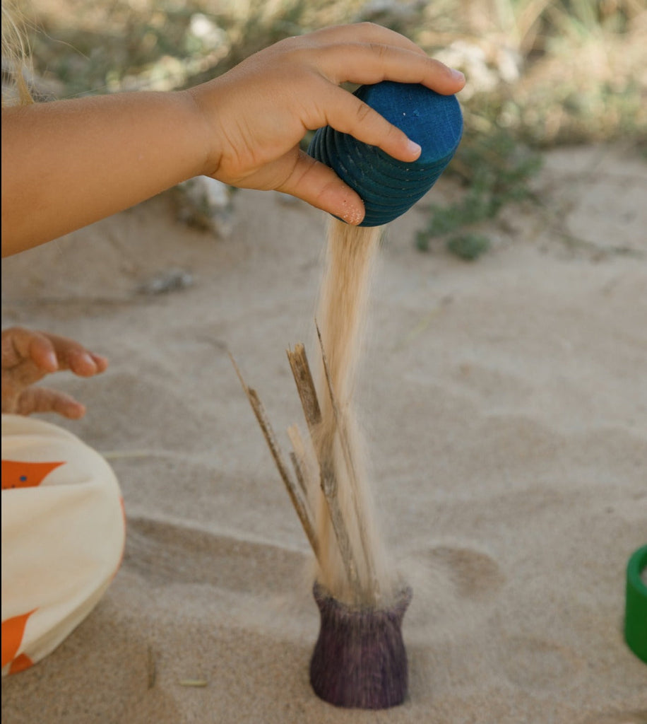 Child dropping sand from a blue Grapat tall wooden pot  into a smalller purple pot at the beach