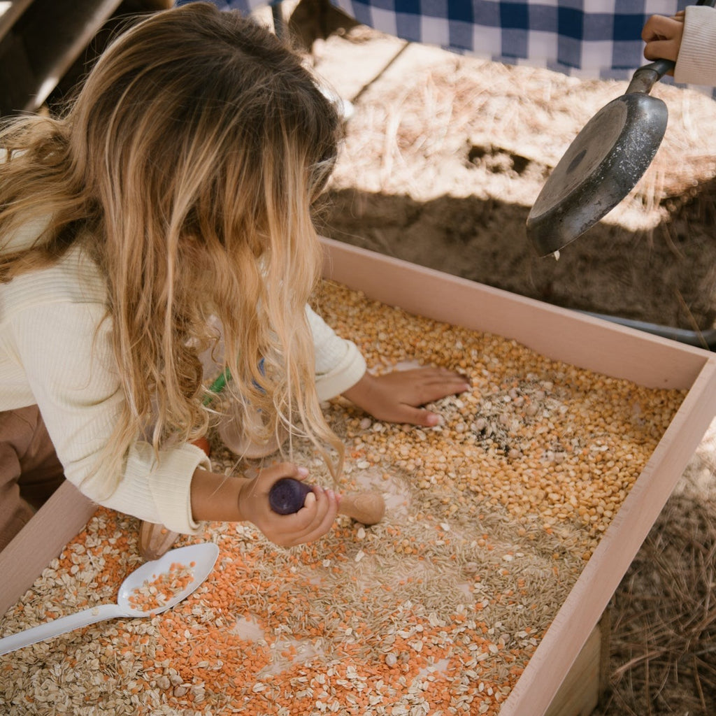 Child using wooden grapat tools in a large wooden tray with sensory food items 