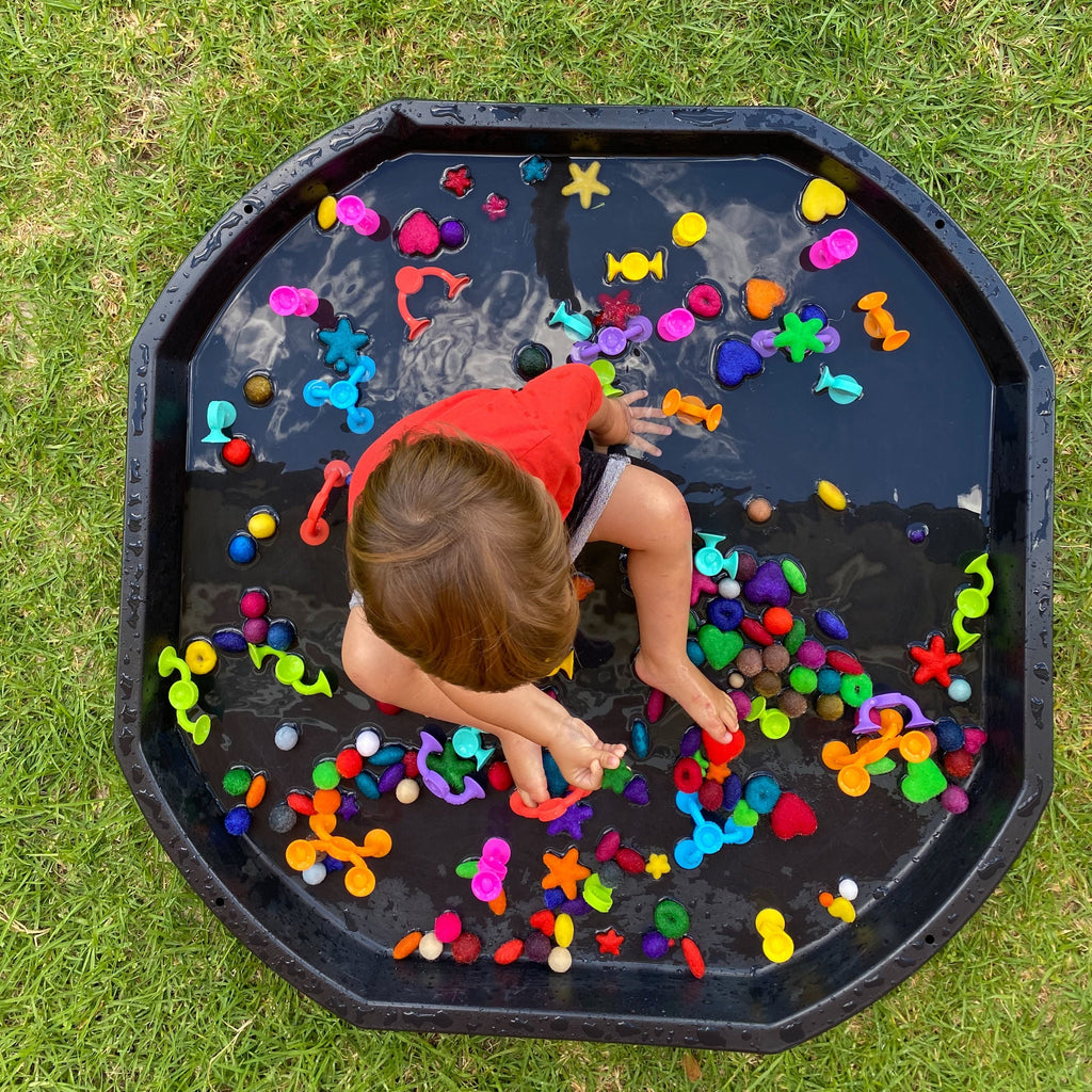 Boy sitting in tuff tray outside which is wet with fat brain toy sensory items and felt balls