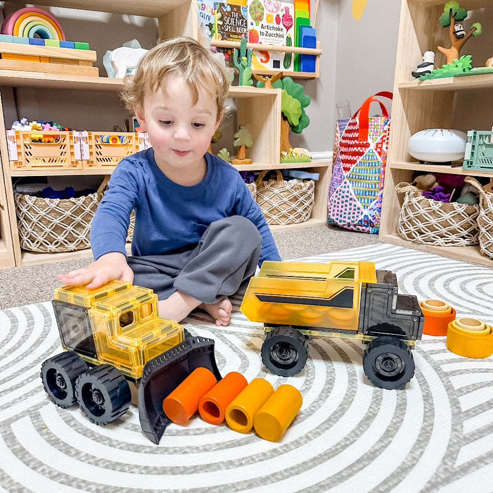 Child playing with magnetic tile toy construction vehicles on a patterned rug in a playroom.