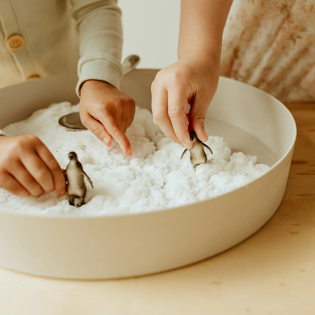 Children playing with toy penguins in a bowl of snow on a wooden surface