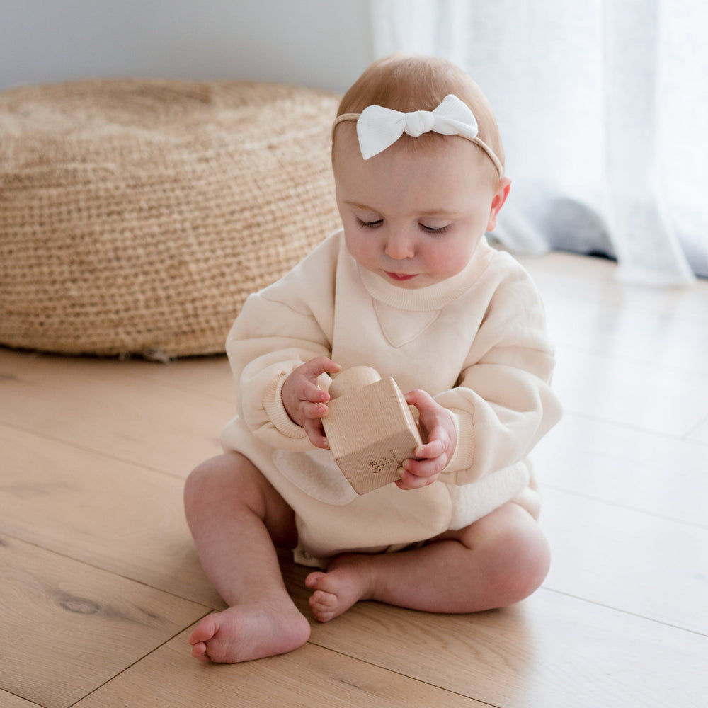 A toddler holding The Pincer Puzzle a montessori wooden toy sitting on ground with a ribbon in hair and wearing cream jersey onesie