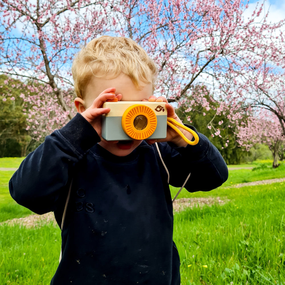 Child taking photo with wooden camera from August Play Subscription box with pink blossoms behind him