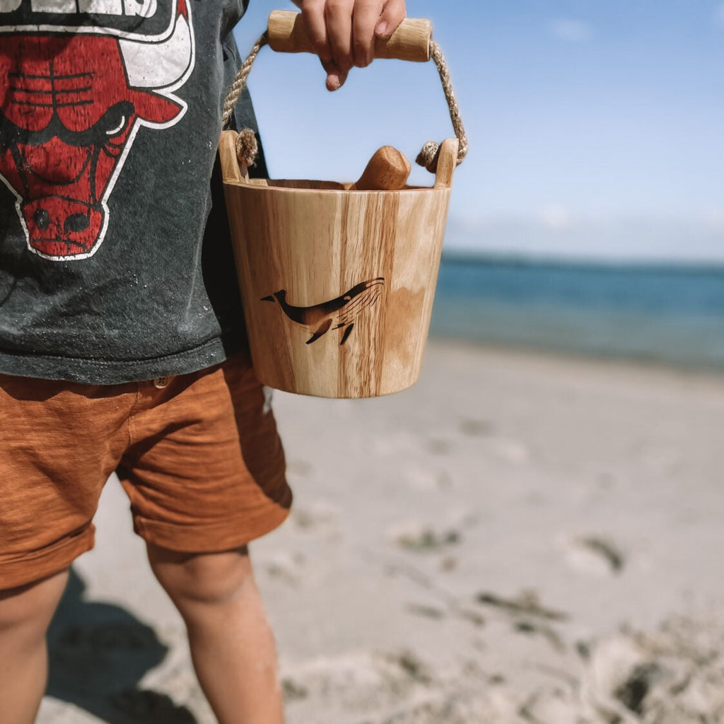 Child in shorts and tshirt holding wooden bucket and scoop with whale burn on front on sandy beach