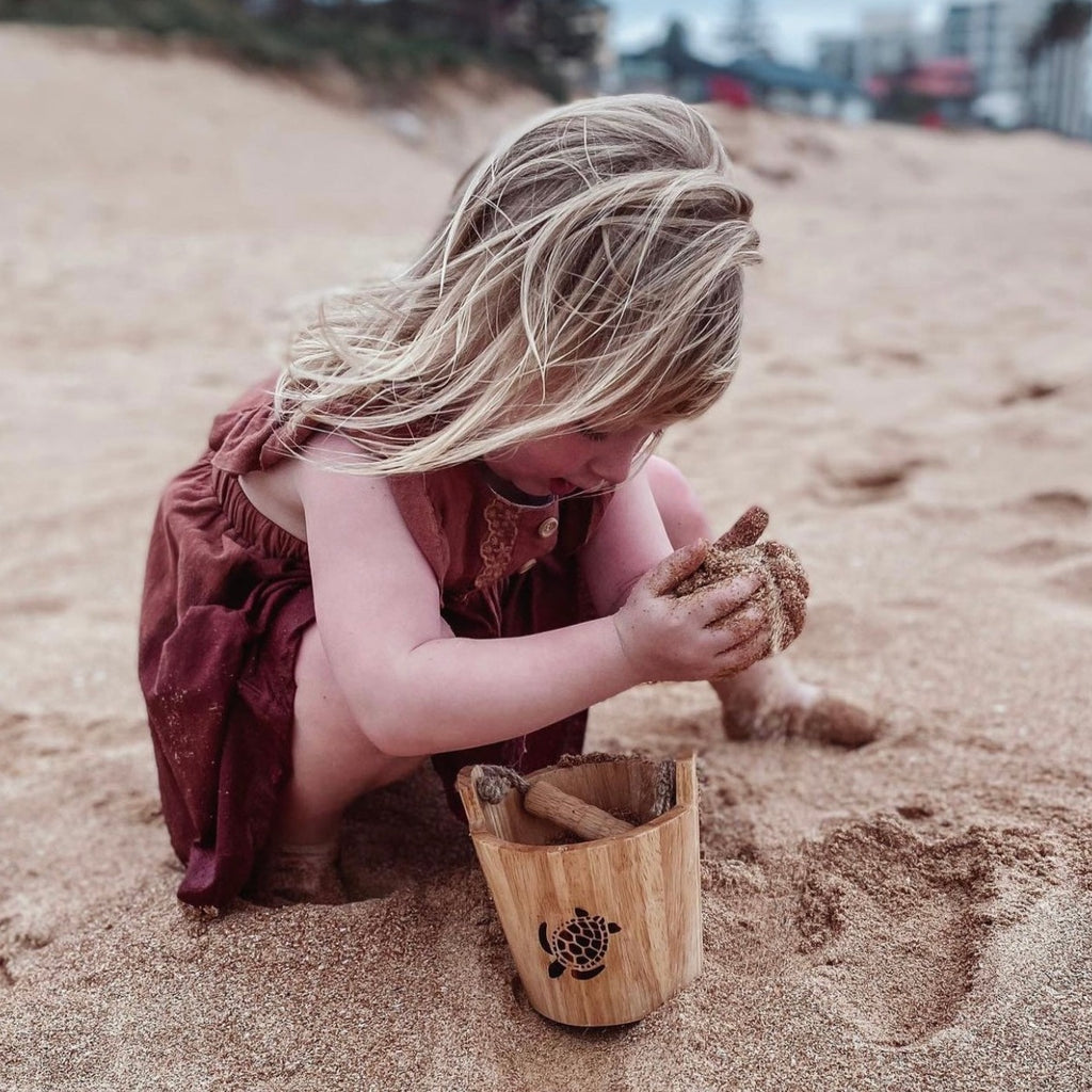 Girl in brown dress holding Explore Nook Wooden Bucket and Scoop set in turtle burn stamp edition on beach