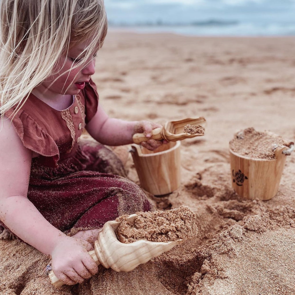 Girl in brown dress holding and playing with Explore Nook Wooden Bucket and Scoop set in turtle burn stamp edition on beach