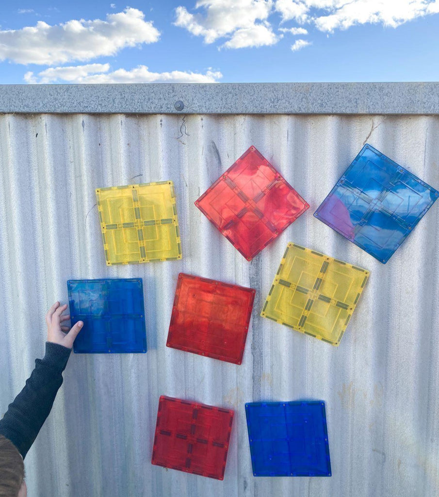 Child holding up large square magnetic tile on corrugated shed with other red, blue, and yellow tiles with blue sky and clouds above